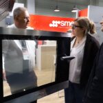 Trzy osoby stoją przed dużym piecem przemysłowym na targach. Jeden z mężczyzn, ubrany w białą koszulę i identyfikator, wyjaśnia funkcje pieca kobiecie w okularach i mężczyźnie w ciemnej kurtce. Drzwi piekarnika są częściowo otwarte. projekt i nadzór Tatjana Kobuszewska trksystem.com