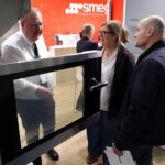 Trzy osoby stoją przed dużym przemysłowym piekarnikiem ze stali nierdzewnej ze szklanymi drzwiczkami na wystawie. Jeden z mężczyzn objaśnia urządzenie kobiecie i mężczyźnie. W tle znajduje się jasnopomarańczowy wyświetlacz Smeg Professional i oznakowanie informacyjne. projekt i nadzór Tatjana Kobuszewska trksystem.com