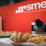 Dwie tace z różnymi rogalikami są wystawione na ladzie przed jaskrawoczerwoną ścianą z logo smeg PROFESSIONAL. W tle widoczna jest kremowa lodówka i mały piekarnik. projekt i nadzór Tatjana Kobuszewska trksystem.com