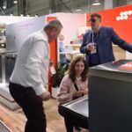 Cztery dorosłe osoby wchodzą w interakcję w salonie z urządzeniami kuchennymi. Mężczyzna w garniturze stoi w pobliżu zmywarki, rozmawiając i trzymając filiżankę. Kobieta klęczy przy otwartej zmywarce, podczas gdy dwóch innych mężczyzn, jeden stojący, a drugi pochylony, obserwuje. Wyświetlane są nowoczesne piekarniki i zlewozmywaki. projekt i nadzór Tatjana Kobuszewska trksystem.com
