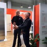 Dwóch mężczyzn stoi w nowoczesnym salonie, rozmawiając o dużej lodówce ze stali nierdzewnej. Jeden z nich gestykuluje w kierunku urządzenia. Otaczają ich wyświetlacze informacyjne, czerwony branding i zielone rośliny. Przestrzeń ma drewnianą podłogę i oświetlenie sufitowe w stylu industrialnym. projekt i nadzór Tatjana Kobuszewska trksystem.com