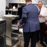 Cztery dorosłe osoby stoją wokół dwóch otwartych komercyjnych zmywarek do naczyń podczas imprezy w pomieszczeniu. Jeden mężczyzna, ubrany w niebieską kurtkę, ładuje naczynia, podczas gdy inni uważnie obserwują. W tle znajdują się stoły, jedzenie i wystawy. projekt i nadzór Tatjana Kobuszewska trksystem.com