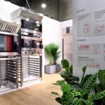 Nowoczesna ekspozycja kuchni komercyjnej w Stoisku Smeg Professional - 50 m² w sąsiedztwie Grafena obejmuje piekarniki i regały ze stali nierdzewnej na drewnianej podłodze. Biała ściana przedstawia polską grafikę informacyjną, a zielone rośliny doniczkowe dodają scenie odrobiny natury. projekt i nadzór Tatjana Kobuszewska trksystem.com