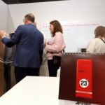 Ludzie oglądają duże metalowe urządzenie na stoisku targowym z białymi ścianami i polskim tekstem. Na pierwszym planie spiralny kalendarz biurkowy z czerwonymi drzwiami i liczbą 23. Cztery dorosłe osoby wchodzą w interakcję z wyświetlaczem w tle. projekt i nadzór Tatjana Kobuszewska trksystem.com