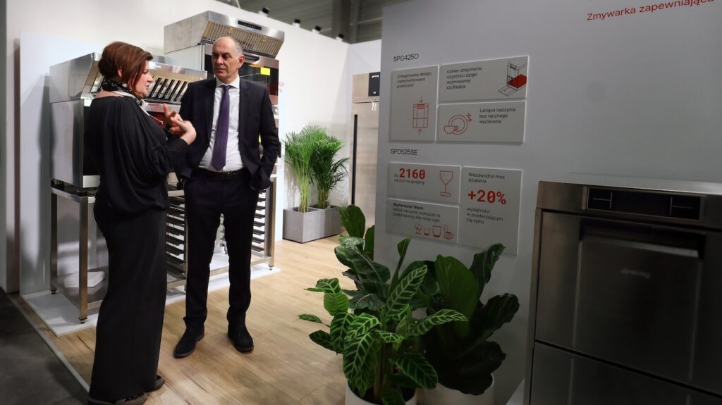 Kobieta i mężczyzna w strojach biznesowych rozmawiają w nowoczesnym salonie z urządzeniami kuchennymi ze stali nierdzewnej i zielonymi roślinami w Stoisku Smeg Professional - 50 m² w sąsiedztwie Grafeny. Na pierwszym planie widoczna jest tablica z polskim tekstem na temat zmywarki. projekt i nadzór Tatjana Kobuszewska trksystem.com