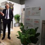 Kobieta i mężczyzna w strojach biznesowych rozmawiają w nowoczesnym salonie z urządzeniami kuchennymi ze stali nierdzewnej i zielonymi roślinami w Stoisku Smeg Professional - 50 m² w sąsiedztwie Grafeny. Na pierwszym planie widoczna jest tablica z polskim tekstem na temat zmywarki. projekt i nadzór Tatjana Kobuszewska trksystem.com