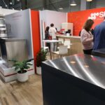 Stoisko Smeg Professional - 50 m² w sąsiedztwie Grafena prezentuje duże urządzenia kuchenne ze stali nierdzewnej, a odwiedzający mogą odkrywać sprzęt wśród roślin doniczkowych, nowoczesnych białych krzeseł i stołów oraz żywych czerwonych ścian z logo SMEG z polskim tekstem. projekt i nadzór Tatjana Kobuszewska trksystem.com