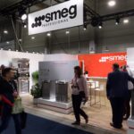 Grupa ludzi przechodzi obok i staje wewnątrz nowoczesnego stoiska wystawowego SMEG Professional, na którym znajdują się urządzenia kuchenne, rośliny i ekspozycje produktów w jasnym świetle, z markowym oznakowaniem i wnętrzem z drewnianą podłogą. projekt i nadzór Tatjana Kobuszewska trksystem.com