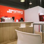 Nowoczesne stoisko wystawowe z czerwoną ścianą SMEG Professional, białą lodówką SMEG w stylu retro, ekspresem do kawy, białymi krzesłami, ladą i stołem z monitorem komputerowym. Drewniana podłoga i industrialne oświetlenie sufitowe dopełniają całości. projekt i nadzór Tatjana Kobuszewska trksystem.com