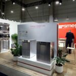 Nowoczesne stoisko targowe dla Smeg Professional obejmuje urządzenia kuchenne ze stali nierdzewnej, białe ściany z czerwonym brandingiem, panele informacyjne, rośliny doniczkowe i mężczyznę w garniturze stojącego przy ladzie, a wszystko to przy jasnym oświetleniu górnym. projekt i nadzór Tatjana Kobuszewska trksystem.com
