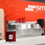 Nowoczesna ekspozycja kuchenna z kremową lodówką SMEG, srebrnym ekspresem do kawy La Pavoni, czerwonymi filiżankami SMEG ułożonymi na górze, ścianą z logo SMEG Professional, małym kalendarzem i sprzętem do kawy na białej ladzie na czerwonym tle. projekt i nadzór Tatjana Kobuszewska trksystem.com