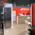 Nowoczesny salon wystawowy z urządzeniami kuchennymi Smeg, w tym lodówkami ze stali nierdzewnej, piekarnikami i maszynami nablatowymi. Przestrzeń ma drewnianą podłogę, białe krzesła przy drewnianej ladzie, jaskrawoczerwony branding, informacyjną grafikę ścienną i zielone rośliny. projekt i nadzór Tatjana Kobuszewska trksystem.com