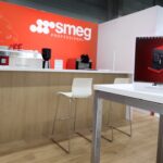 Nowoczesny salon kuchenny z białą lodówką SMEG, czerwonymi ekspresami do kawy SMEG na ladzie, białymi stołkami i stołem z czerwonym kalendarzem SMEG. Tło ściany jest jaskrawoczerwone z logo SMEG i napisem PROFESSIONAL. projekt i nadzór Tatjana Kobuszewska trksystem.com