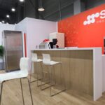 Nowoczesny salon wystawowy z ekspozycją Smeg Professional ma czerwono-białą ścianę, ladę barową z białymi stołkami, wbudowane urządzenia kuchenne, lodówkę ze stali nierdzewnej, piekarnik i drewnianą podłogę pod jasnym oświetleniem sufitowym. projekt i nadzór Tatjana Kobuszewska trksystem.com