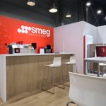 Nowoczesny showroom z podłogą z jasnego drewna, białymi stołami i krzesłami, kremową lodówką Smeg w stylu retro, ekspresem do kawy i czerwoną ścianą Smeg Professional z ilustracjami narzędzi w tle. Jasne oświetlenie i czysty, minimalistyczny design. projekt i nadzór Tatjana Kobuszewska trksystem.com