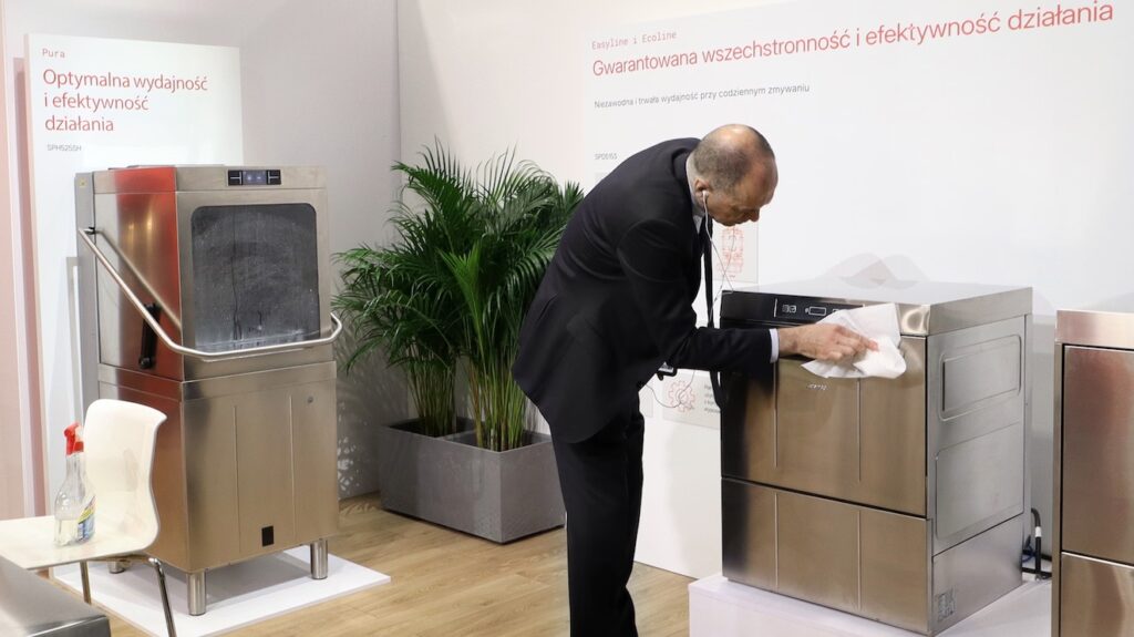 W stoisku Smeg Professional - 50 m² w sąsiedztwie Grafeny, mężczyzna w garniturze poleruje zmywarkę ze stali nierdzewnej na wystawie, a obok znajduje się inna zmywarka, butelka z rozpylaczem, rośliny doniczkowe i polskie plakaty informacyjne. projekt i nadzór Tatjana Kobuszewska trksystem.com