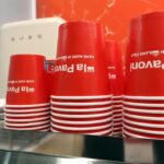 Stos czerwonych papierowych filiżanek do kawy z białymi brzegami i tekstem "Hand Made in Italy" do góry nogami, umieszczonych na białych spodkach. Tło obejmuje rozmytą czerwoną ścianę i część białego urządzenia. projekt i nadzór Tatjana Kobuszewska trksystem.com
