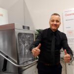 Mężczyzna w czarnej koszuli uśmiecha się i pokazuje dwa kciuki w górę obok dużej srebrnej zmywarki. Za nim biało-pomarańczowe napisy w języku polskim mówią o wydajności i niezawodności. Otoczenie wygląda na nowoczesny salon wystawowy lub stoisko wystawowe. projekt i nadzór Tatjana Kobuszewska trksystem.com