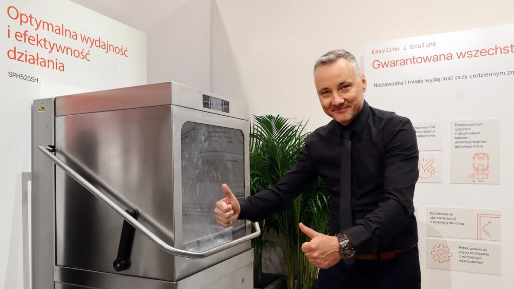 Uśmiechnięty mężczyzna w czarnej koszuli stoi obok przemysłowej zmywarki, trzymając dwa kciuki w górę na Stoisku Smeg Professional - 50 m² w sąsiedztwie Grafen. Za nim polski tekst i zielone rośliny zdobią strefę demonstracyjną produktu. projekt i nadzór Tatjana Kobuszewska trksystem.com
