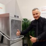 Uśmiechnięty mężczyzna w czarnej koszuli stoi obok przemysłowej zmywarki, trzymając dwa kciuki w górę na Stoisku Smeg Professional - 50 m² w sąsiedztwie Grafen. Za nim polski tekst i zielone rośliny zdobią strefę demonstracyjną produktu. projekt i nadzór Tatjana Kobuszewska trksystem.com
