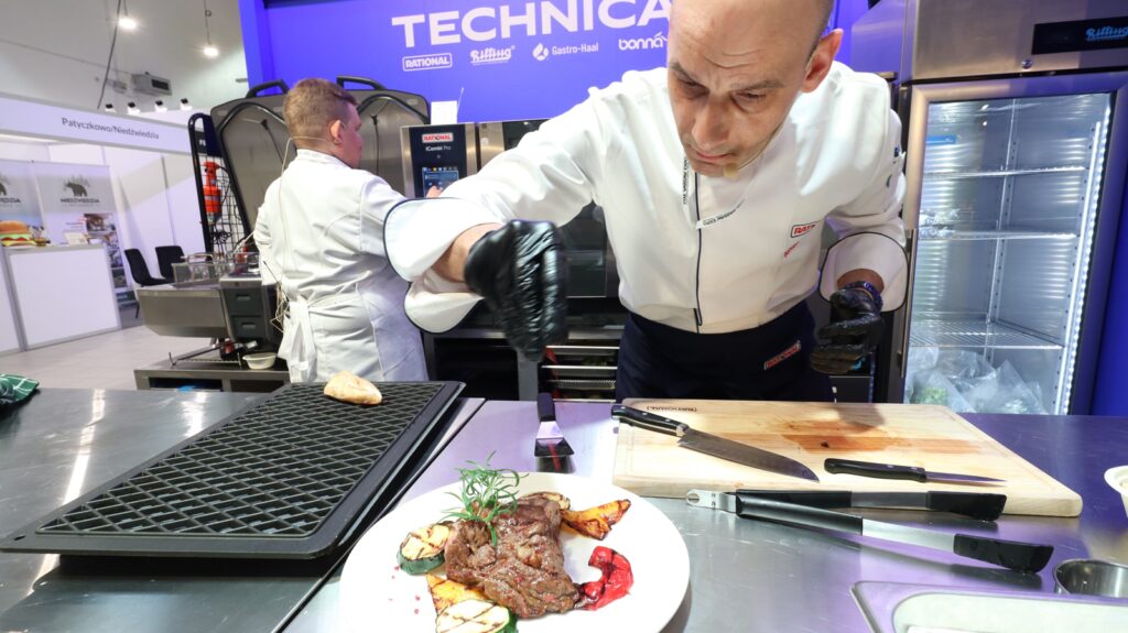 Szef kuchni w białym fartuchu i czarnych rękawiczkach nalewa sos na stek przyozdobiony ziołami i warzywami. Inny kucharz pracuje w tle w strefie pokazów gotowania Stoisko Technica Eurogastro z widocznymi oznakowaniami technicznymi. projekt i nadzór Tatjana Kobuszewska trksystem.com