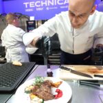 Szef kuchni w białym fartuchu i czarnych rękawiczkach nalewa sos na stek przyozdobiony ziołami i warzywami. Inny kucharz pracuje w tle w strefie pokazów gotowania Stoisko Technica Eurogastro z widocznymi oznakowaniami technicznymi. projekt i nadzór Tatjana Kobuszewska trksystem.com