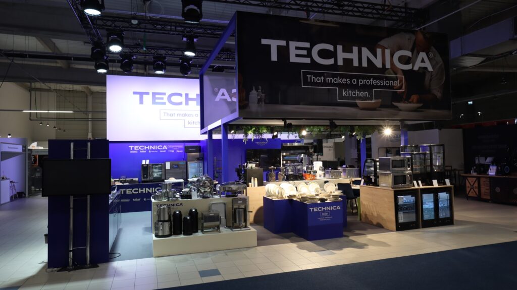 Pokazano nowoczesne stoisko wystawiennicze "TECHNICA" z dużymi znakami, urządzeniami kuchennymi i sprzętem na wystawie. Stoisko jest oświetlone reflektorami i udekorowane na biało i niebiesko, podkreślając profesjonalną technologię kuchenną. projekt i nadzór Tatjana Kobuszewska trksystem.com