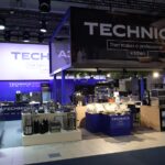 Pokazano nowoczesne stoisko wystawiennicze "TECHNICA" z dużymi znakami, urządzeniami kuchennymi i sprzętem na wystawie. Stoisko jest oświetlone reflektorami i udekorowane na biało i niebiesko, podkreślając profesjonalną technologię kuchenną. projekt i nadzór Tatjana Kobuszewska trksystem.com