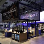 Nowoczesne stoisko wystawowe prezentuje urządzenia kuchenne, takie jak piekarniki, lodówki i kuchenki. Stoisko jest niebiesko-białe, z logo "TECHNICA" na dużych znakach nad i wokół ekspozycji. Kilka zestawów kuchennych i miejsc do siedzenia jest widocznych przy jasnym oświetleniu. projekt i nadzór Tatjana Kobuszewska trksystem.com