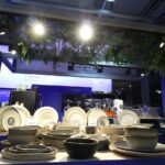 Ekspozycja beżowych ceramicznych talerzy i misek stoi na ladzie pod jasnym światłem w Stoisku Technica na Eurogastro w Ptak Warsaw Expo - 70 m² z niebiesko-białym brandingiem. Powyżej wiszą zielone rośliny liściaste, ustawione na świecącym niebieskim tle. projekt i nadzór Tatjana Kobuszewska trksystem.com