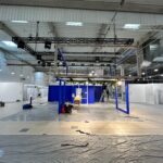 Duża przestrzeń wewnętrzna w budowie z metalową konstrukcją ramową pośrodku, otoczona narzędziami, sprzętem i plastikowymi arkuszami na podłodze. Tło stanowią niebieskie panele i białe ściany, a z sufitu zwisają światła i kable. projekt i nadzór Tatjana Kobuszewska trksystem.com