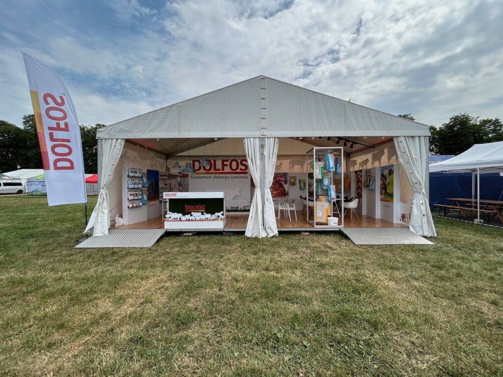 Biały namiot eventowy z otwartymi bokami wyświetla znak "DOLFOS" nad wejściem. Wewnątrz znajdują się półki z produktami, banery i broszury. Namiot stoi na trawie, a wysoka flaga z napisem "DOLFOS" stoi po lewej stronie pod częściowo zachmurzonym niebem. projekt i nadzór Tatjana Kobuszewska trksystem.com