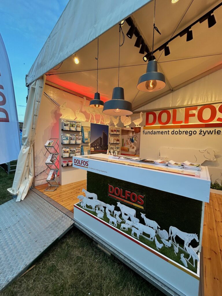 Stoisko targowe z białym namiotem, drewnianą podłogą i brandingiem DOLFOS. Lada wyświetla broszury i ma sylwetki zwierząt hodowlanych. Broszury i plakaty są widoczne na ścianach, a nad ladą wiszą trzy czarne lampy wiszące. projekt i nadzór Tatjana Kobuszewska trksystem.com