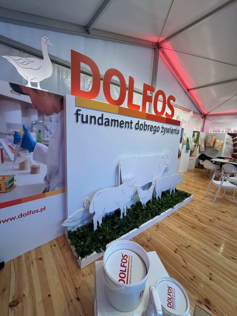 Stoisko targowe z napisem DOLFOS fundament dobrego żywienia, przedstawiające sylwetki zwierząt (krowy, owce, kozy) na białym tle z zielonymi liśćmi, wanny z napisem DOLFOS oraz nowoczesne, jasne wnętrze z drewnianą podłogą. projekt i nadzór Tatjana Kobuszewska trksystem.com