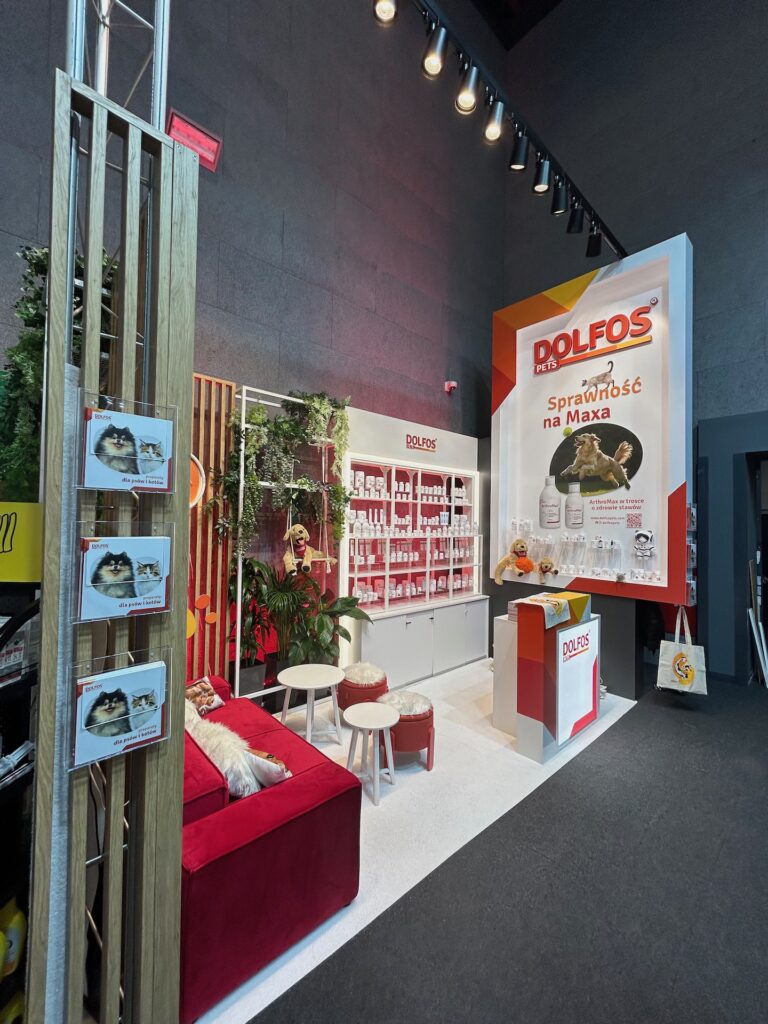 Stoisko targowe firmy Dolfos ma czerwono-białą kolorystykę, czerwoną sofę, okrągłe stoły, półki z produktami dla zwierząt oraz duży baner przedstawiający suplementy dla zwierząt i psa, z napisem "Dolfos" i "Sprawność na Maxa". projekt i nadzór Tatjana Kobuszewska