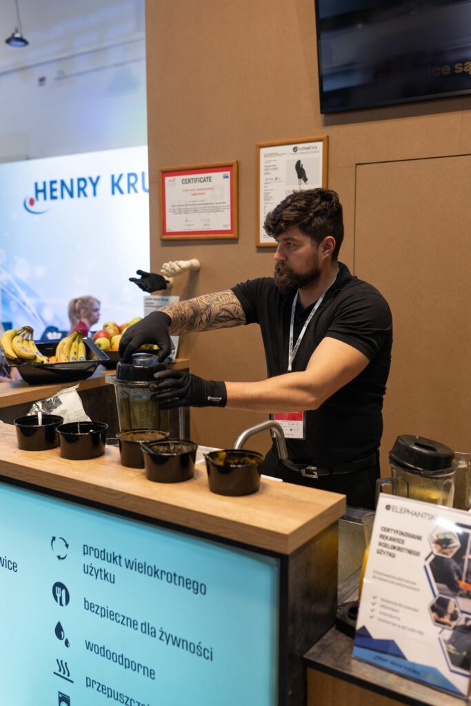 Brodaty mężczyzna o ciemnych włosach i z tatuażem przygotowuje smoothie przy ladzie, używając blendera. Ma na sobie czarne rękawiczki i czarną koszulkę. W pobliżu leżą banany i miski do przygotowywania składników. Na ścianie i ladzie widoczne są tabliczki i certyfikaty, w tym te związane z projektem i nadzorem Tatjany Kobuszewskiej, co sugeruje profesjonalne kwalifikacje osób obsługujących stoisko targowe w. projekt i nadzór Tatjana Kobuszewska