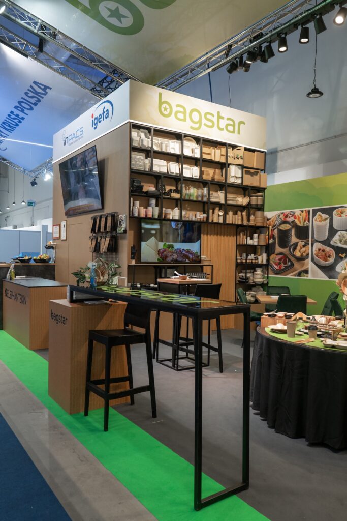 Stoisko targowe Bagstar i Henry Kruse na Eurogastro 2025 w Ptak Warsaw Expo prezentuje ekologiczne opakowania z zielonymi i drewnianymi akcentami, otwarty tył stoiska oraz kartonową rakietę jako akcent wśród kubków i miseczek na próbki. Stoisko Bagstar i Henry Kruse na Eurogastro 2025 w Ptak Warsaw Expo, 100 m². Pierwsza wspólna realizacja w grupie igefa. Bagstar: zielona paleta, brązowa recepcja symbol materiałów z odzysku, kartonowa rakieta jako akcent. Henry Kruse: chłodna biało-błękitna paleta. Otwarty tył stoiska. Projekt Tatjany Kobuszewskiej, TRK System.
