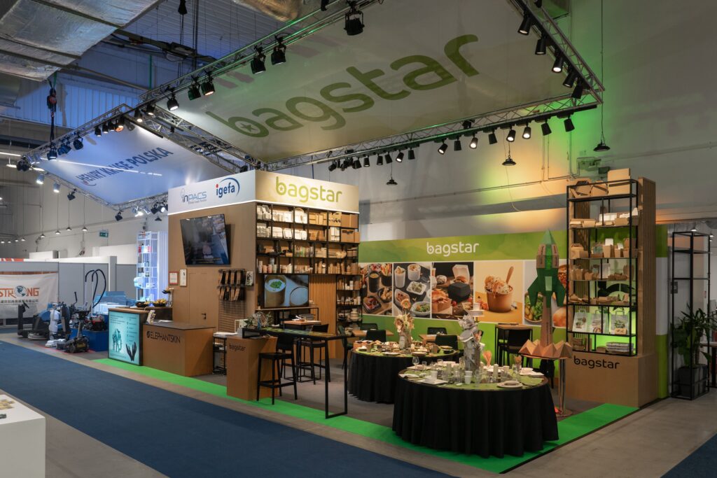 Stoisko targowe firmy "bagstar" prezentuje różne ekologiczne produkty opakowaniowe. Półki wypełnione są torbami i pojemnikami. W nowoczesnej aranżacji znajduje się okrągły stół z próbkami, ekran, banery, zielone oświetlenie i oznakowanie marki. Stoisko Bagstar i Henry Kruse na Eurogastro 2025 w Ptak Warsaw Expo, 100 m². Pierwsza wspólna realizacja w grupie igefa. Bagstar: zielona paleta, brązowa recepcja symbol materiałów z odzysku, kartonowa rakieta jako akcent. Henry Kruse: chłodna biało-błękitna paleta. Otwarty tył stoiska. Projekt Tatjany Kobuszewskiej, TRK System.