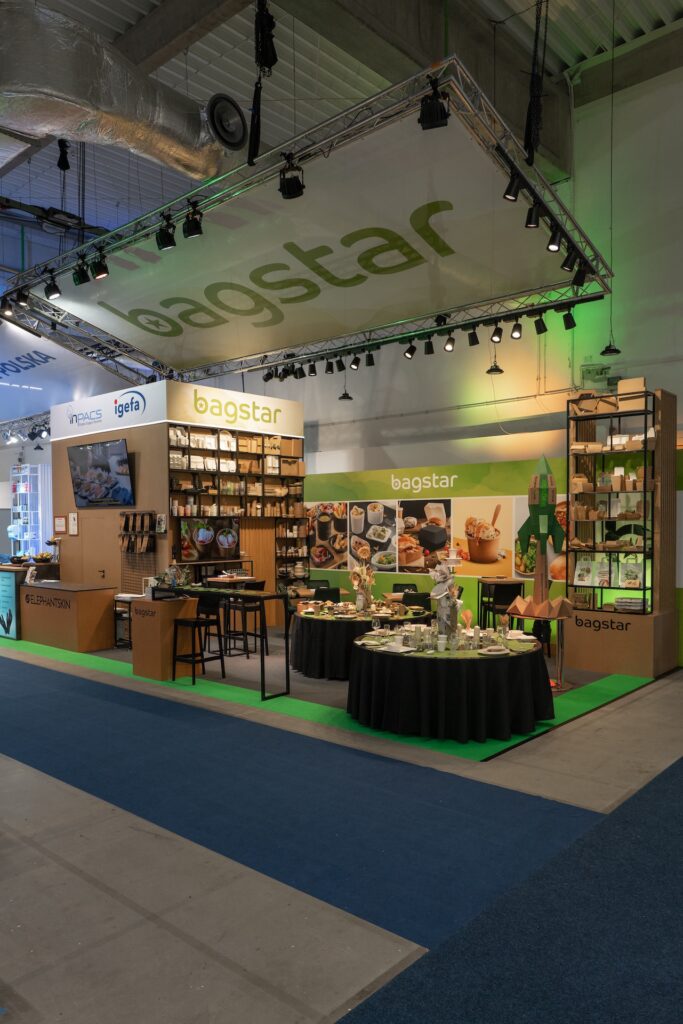Stoisko targowe firmy Bagstar prezentuje ekologiczne produkty opakowaniowe. Na półkach znajdują się brązowe torby, pudełka i kubki. Stoły prezentują artykuły spożywcze i próbki opakowań. Jasne oświetlenie, zielone akcenty i oznakowanie marki są widoczne w całej nowoczesnej konfiguracji. Stoisko Bagstar i Henry Kruse na Eurogastro 2025 w Ptak Warsaw Expo, 100 m². Pierwsza wspólna realizacja w grupie igefa. Bagstar: zielona paleta, brązowa recepcja symbol materiałów z odzysku, kartonowa rakieta jako akcent. Henry Kruse: chłodna biało-błękitna paleta. Otwarty tył stoiska. Projekt Tatjany Kobuszewskiej, TRK System.