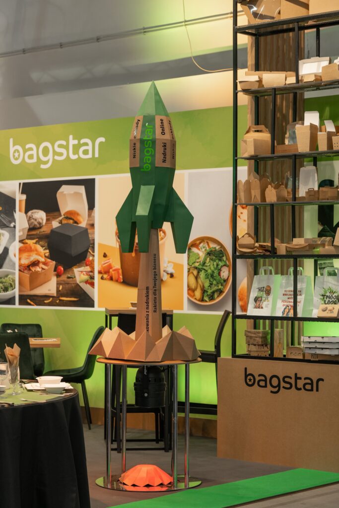 Tekturowa rakieta z brandingiem "bagstar" stoi przed zielono-żółtym stoiskiem z ekologicznymi opakowaniami na żywność. Na półkach znajdują się papierowe torby i pudełka na wynos. W tle wyświetlane są zdjęcia żywności, a na ścianie stoiska widnieje napis "bagstar". Stoisko Bagstar i Henry Kruse na Eurogastro 2025 w Ptak Warsaw Expo, 100 m². Pierwsza wspólna realizacja w grupie igefa. Bagstar: zielona paleta, brązowa recepcja symbol materiałów z odzysku, kartonowa rakieta jako akcent. Henry Kruse: chłodna biało-błękitna paleta. Otwarty tył stoiska. Projekt Tatjany Kobuszewskiej, TRK System.