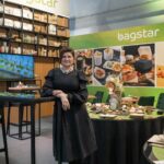 Tatjana Kobuszewska w czarnej sukience stoi przy wysokim stole na stoisku Bagstar i Henry Kruse na Eurogastro 2025 o powierzchni 100 m², zaprojektowanym przez Tatjanę Kobuszewską, charakteryzującym się otwartym układem pleców i zieloną paletą z kartonową rakietą jako akcentem. projekt i nadzór Tatjana Kobuszewska trksystem.com