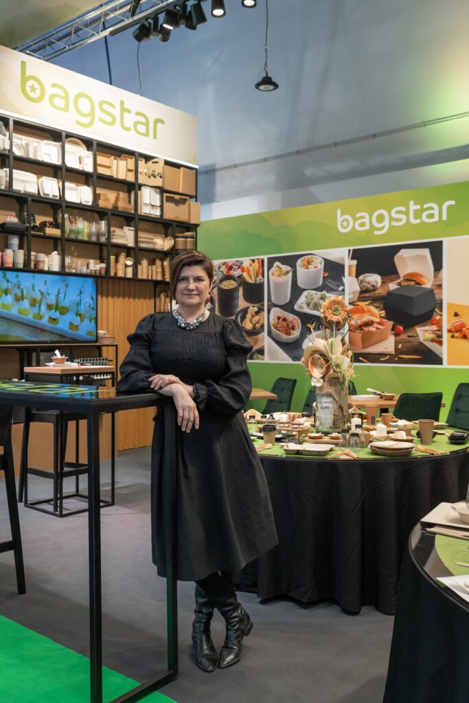 Tatjana Kobuszewska w czarnej sukience stoi przy wysokim stole na stoisku Bagstar i Henry Kruse na Eurogastro 2025 o powierzchni 100 m², zaprojektowanym przez Tatjanę Kobuszewską, charakteryzującym się otwartym układem pleców i zieloną paletą z kartonową rakietą jako akcentem. projekt i nadzór Tatjana Kobuszewska trksystem.com