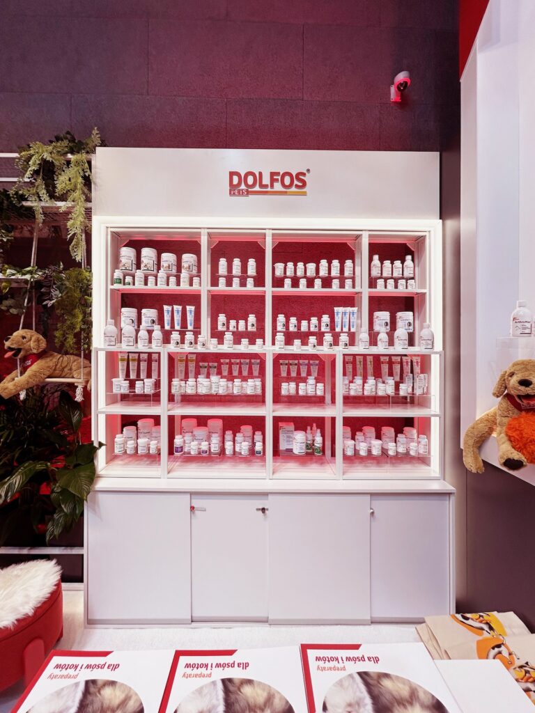 Dobrze oświetlona półka ekspozycyjna wypełniona różnymi produktami do pielęgnacji zwierząt DOLFOS, w tym butelkami, słoikami i tubkami. Półki ekspozycyjne z suplementami Dolfos Pets dla psów i kotów wyróżniają się na tle białej półki z czerwonymi akcentami i pluszowymi zabawkami dla psów po obu stronach. projekt i nadzór Tatjana Kobuszewska