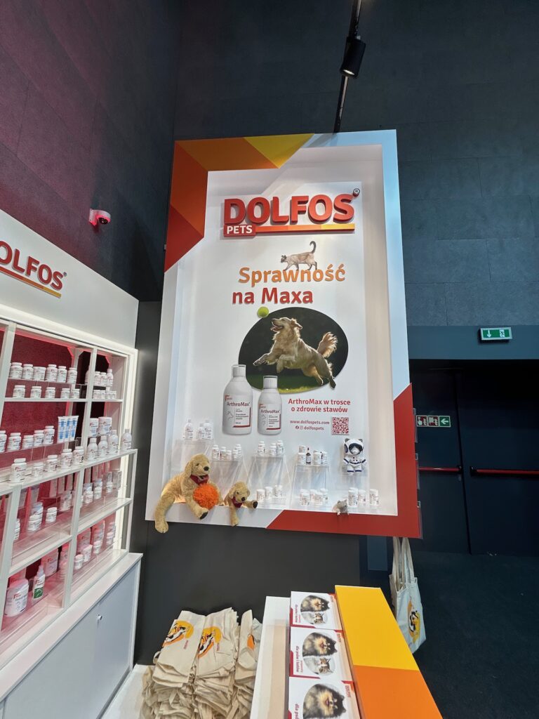 Duża ekspozycja Dolfos Pets prezentuje butelki i słoiki z suplementami dla zwierząt, pluszową zabawkę dla psa i płócienne torby. Branding marki Dolfos Pets na stoisku konferencyjnym świeci czerwono-białym znakiem przedstawiającym skaczącego psa i butelki z suplementami. projekt i nadzór Tatjana Kobuszewska
