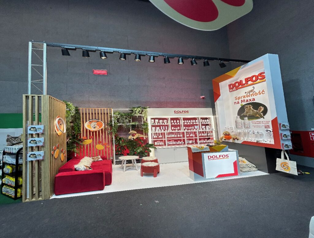 Stoisko reklamowe Dolfos Pets na Międzynarodowym Kongresie Medycyny Weterynaryjnej Małych Zwierząt w Hilton Łódź. Stoisko targowe firmy Dolfos jest utrzymane w biało-czerwonej kolorystyce, z czerwoną kanapą, dekoracjami roślinnymi i półkami na produkty - idealne mobilne stoisko reklamowe. Duży baner z brandingiem Dolfos i polskim tekstem jest zamontowany na prawej ścianie. Projekt Tatjany Kobuszewskiej, TRK System.
