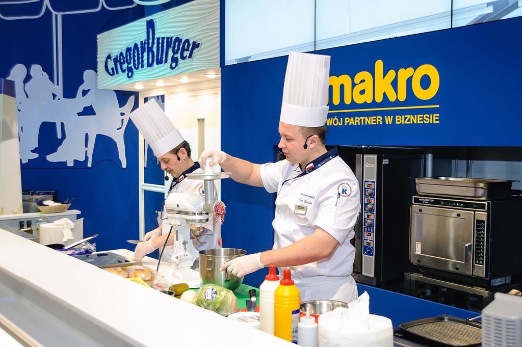 Stoisko targowe live cooking Makro na Eurogastro — strefa pokazów kulinarnych i ekspozycja sprzętu gastronomicznego. Projekt Tatjany Kobuszewskiej, TRK System. Dwóch kucharzy w białych uniformach i wysokich czapkach pracuje przy blacie kuchennym z mikserami i składnikami. Na ladzie znajdują się sosy i produkty. Za nimi na tle niebieskiej ściany widoczne są logo GregoryBurger i makro. W tle widoczny jest również sprzęt kuchenny. Stoisko Bagstar i Henry Kruse na Eurogastro 2025 w Ptak Warsaw Expo, 100 m². Pierwsza wspólna realizacja w grupie igefa. Bagstar: zielona paleta, brązowa recepcja symbol materiałów z odzysku, kartonowa rakieta jako akcent. Henry Kruse: chłodna biało-błękitna paleta. Otwarty tył stoiska. Projekt Tatjany Kobuszewskiej, TRK System.