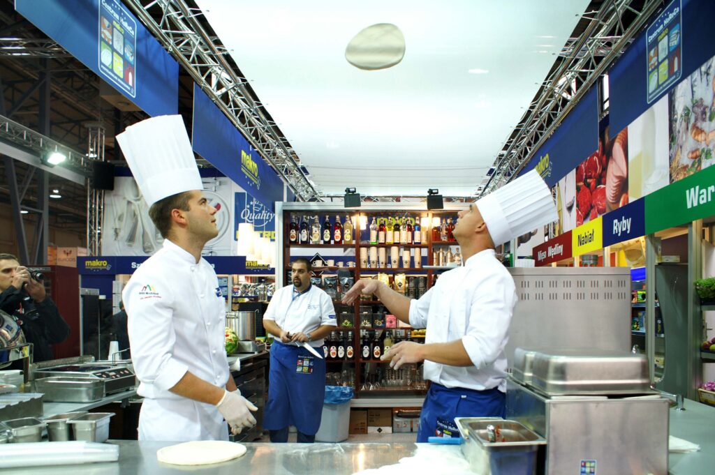 Stoisko targowe live cooking Makro na Polagra — warsztaty kulinarne i część sommelierska, nagrodzone Acanthus Aureus. Projekt Tatjany Kobuszewskiej, TRK System. Dwóch kucharzy w wysokich białych czapkach i uniformach stoi w komercyjnej kuchni. Jeden z nich podrzuca ciasto na pizzę, podczas gdy drugi się temu przygląda. Trzecia osoba pracuje w tle. Kuchnia jest jasna, a półki są wypełnione butelkami i składnikami. projekt i nadzór Tatjana Kobuszewska