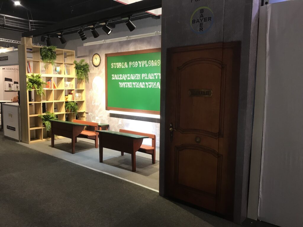 Stoisko konferencyjne Bayer na konferencji lekarzy weterynarii — profesjonalna zabudowa w przestrzeni konferencyjnej. Projekt Tatjany Kobuszewskiej, TRK System
stoisko bayer, zaprojektowanej jak sala lekcyjna, z drewnianymi biurkami, zieloną tablicą do pisania kredą, regałami z roślinami i zegarem. Nad drzwiami widnieje napis SZKOLNA i okrągłe logo Bayer. Tablica kredowa ma polskie pismo z białymi literami. projekt i nadzór Tatjana Kobuszewska