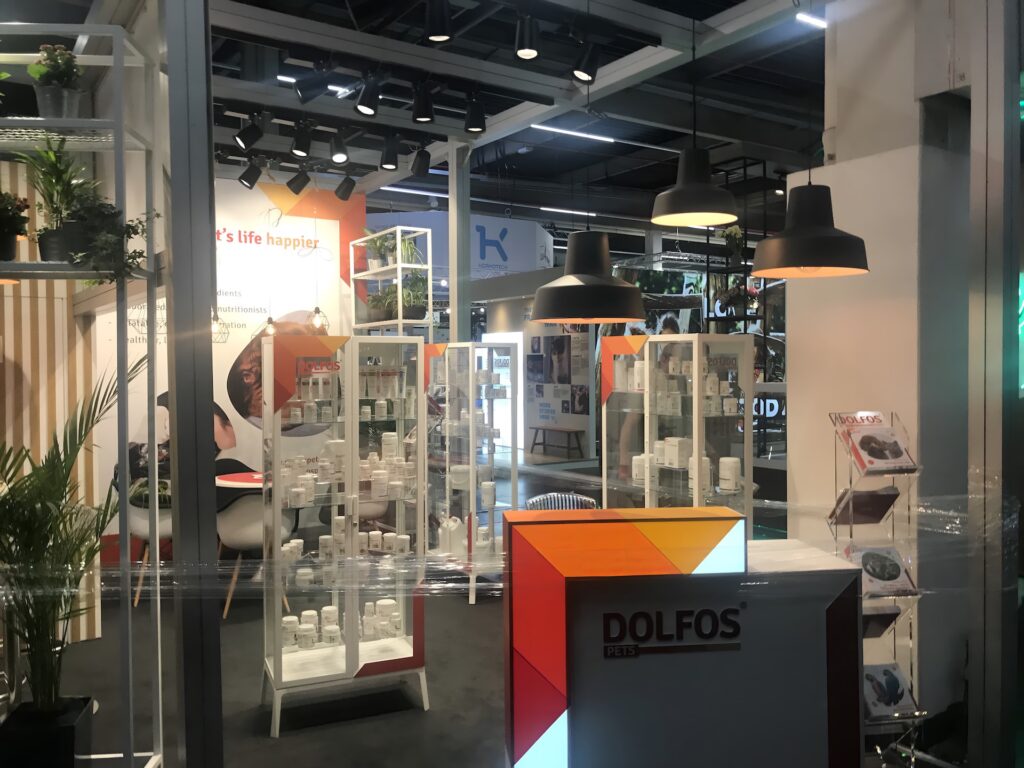 Nowoczesne stoisko Dolfos Pets na targach Interzoo Norymberga obejmuje półki z białymi butelkami produktów, ladę z napisem "DOLFOS", odważne pomarańczowe i żółte akcenty, wiszące czarne lampy oraz grafikę ścienną "pet's life happier". Inne stoiska pojawiają się w tle. projekt i nadzór Tatjana Kobuszewska