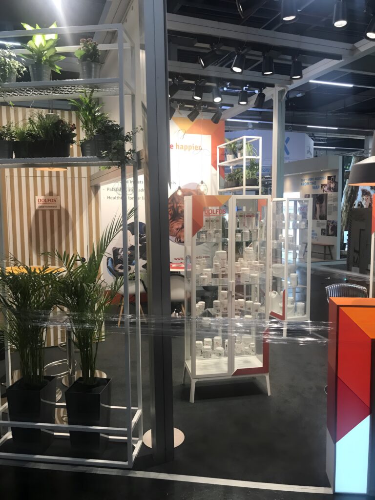 Stoisko dolfos pets na targach Interzoo Norymberga obejmuje wewnętrzne stoisko wystawowe z roślinami doniczkowymi na białych półkach, gablotami z białymi pojemnikami, kolorowymi oznakowaniami, nowoczesnym wystrojem, czarną podłogą, jasnym oświetleniem i plastikową folią rozciągniętą wzdłuż wejścia. projekt i nadzór Tatjana Kobuszewska